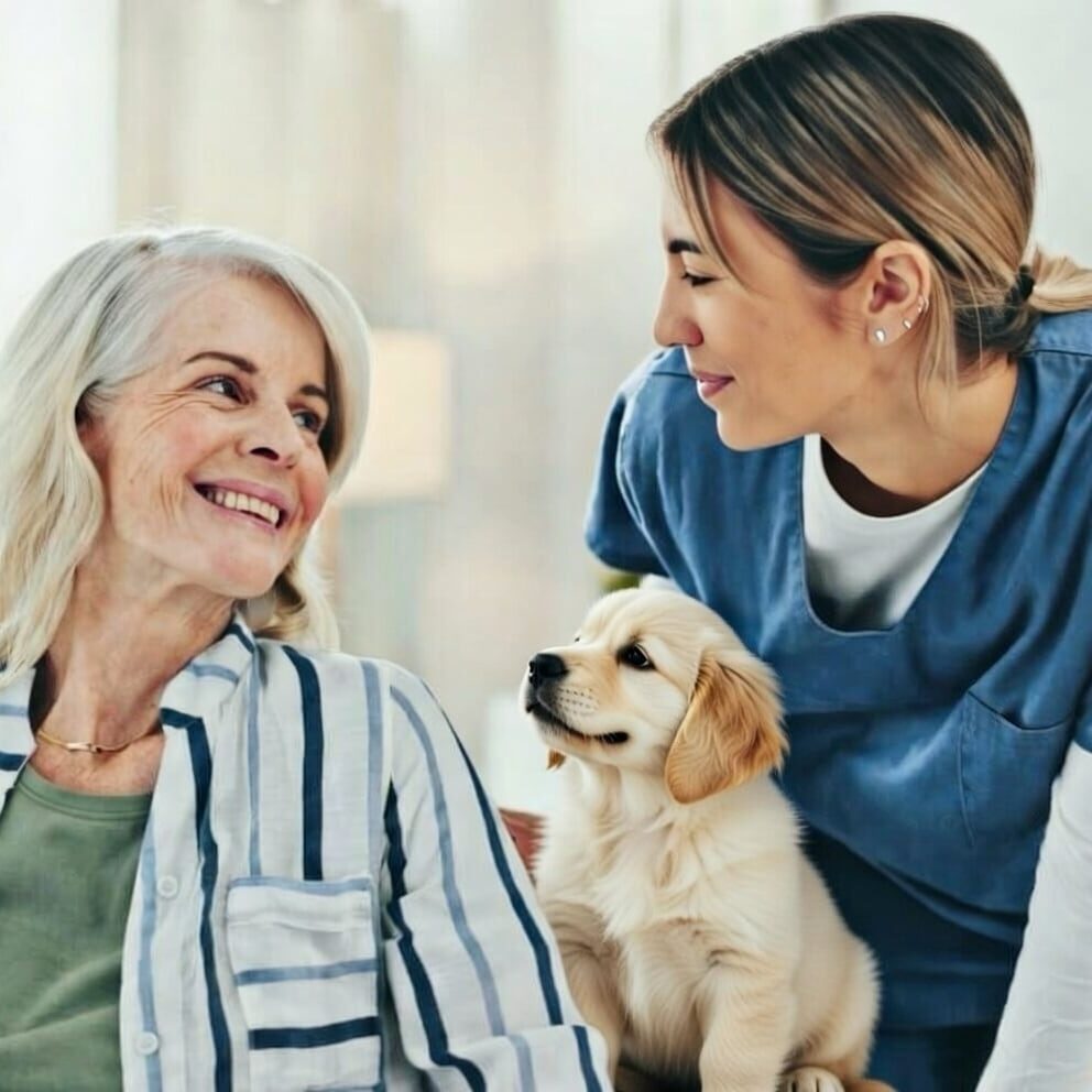An older woman smiles at a younger woman in scrubs who is leaning toward her. A golden retriever puppy sits between them, looking up. The scene appears warm and friendly, suggesting care or companionship.